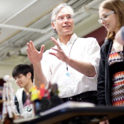 A man in a white button-up gestures to molecular art on a table, explaining it to onlooking students. 