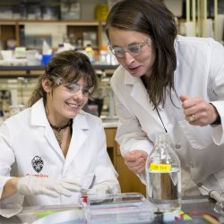 Two women in lab coats work with a sample in a tube in a laboratory.