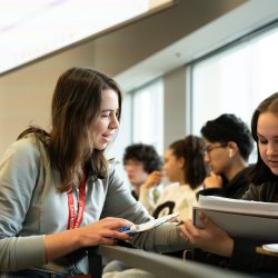 A woman leans over seat railing to aid a student, pointing to the student's notebook mid-conversation.