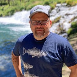 Jason Podrabsky in cap and fish shirt smiling in front of river