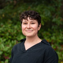 Sophia Bailey smiles in front of Oregon greenery. She wears a black button-up shirt and has short cropped, curly, dark hair.