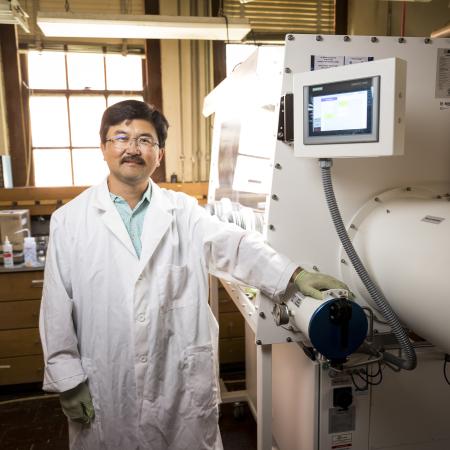 A man in a lab coat stands in front of a white machine used for battery science.