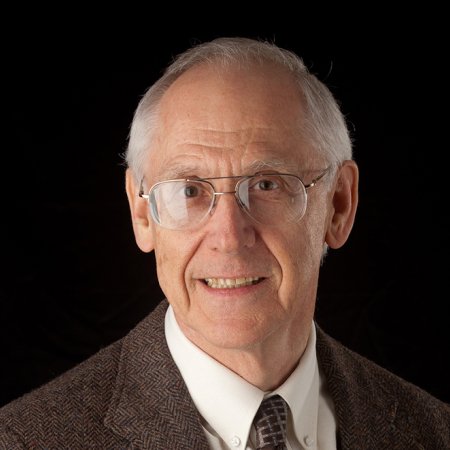 A man's headshot against a black background. The man has gray hair and wears glasses with a brown suit jacket and tie.