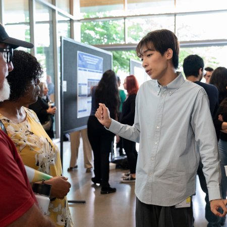 In a busy, sun-lit row lined with research posters, presenters and attendees, a young man in a button-up shirt discusses his poster with two others. 