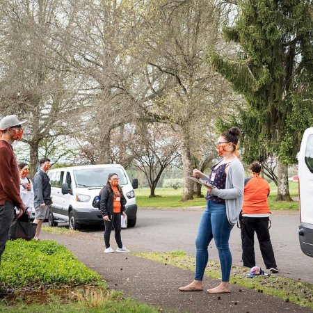 TRACE staff wearing face masks and face shields brief community volunteers before COVID-19 testing outside near two white vans.