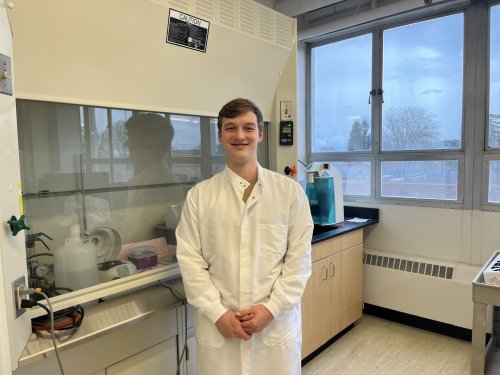 Kelly Shannon dons a white lab coat in an OSU laboratory, equipment lining the wall on his right and a window framing trees outside on his left.