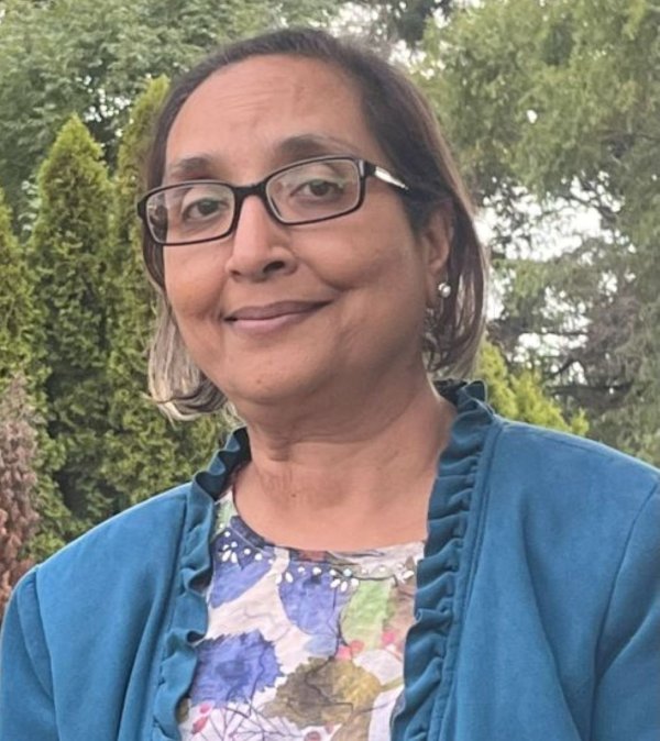 Person wearing glasses and a blue jacket over a floral-patterned top, standing outdoors in front of dense green trees and shrubs in a garden-like setting.