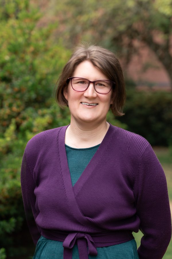 Lauren Dalton smiles outside in a purple wrap sweater. She wears glasses and has short, brown hair.