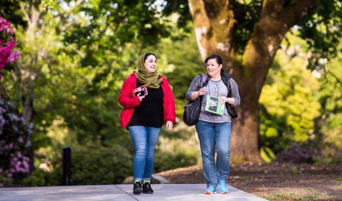 Two female students walking through campus in front of tree
