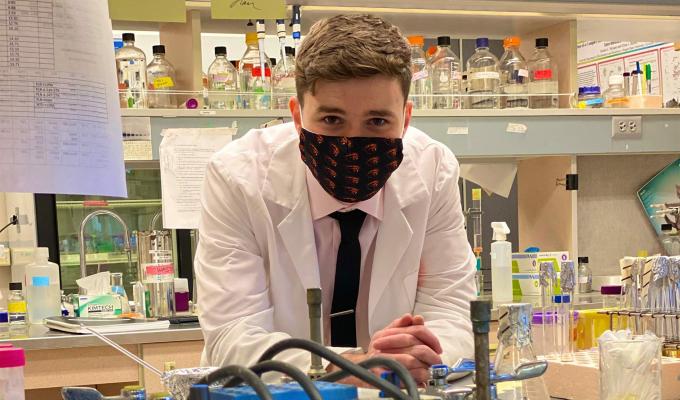 A young man in a lab coat and face mask leaning over a counter with test tubs