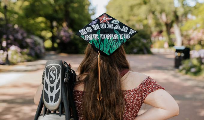 Simone Burton facing away from the camera, holding her scuba fins and wearing her decorated grad cap that says "Honorary Mermaid".