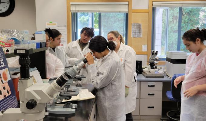 High school students stand around a microscope in a lab wearing lab coats.