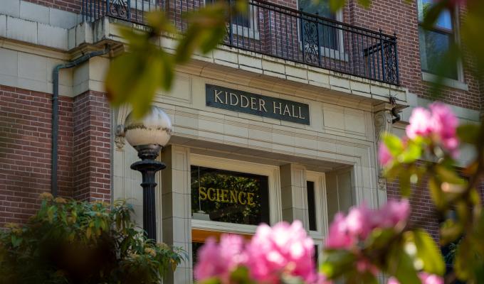 Photo of Kidder Hall from a low angle looking at a door with science written in the glass above the door, and Kidder Hall written in the stone above the glass. Blurred pink flowers appear in the foreground.