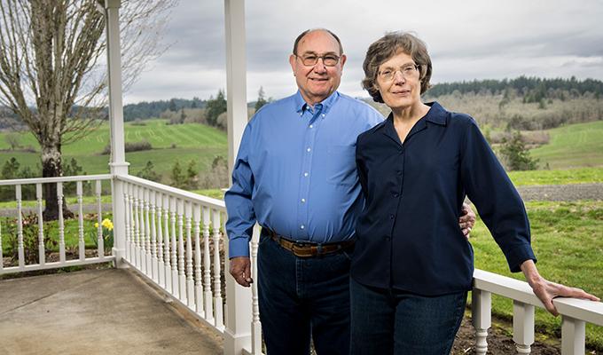 Norbert and Eileen Hartmann lean against a porch railing with green landscape behind them.