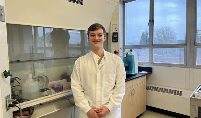 Kelly Shannon dons a white lab coat in an OSU laboratory, equipment lining the wall on his right and a window framing trees outside on his left.
