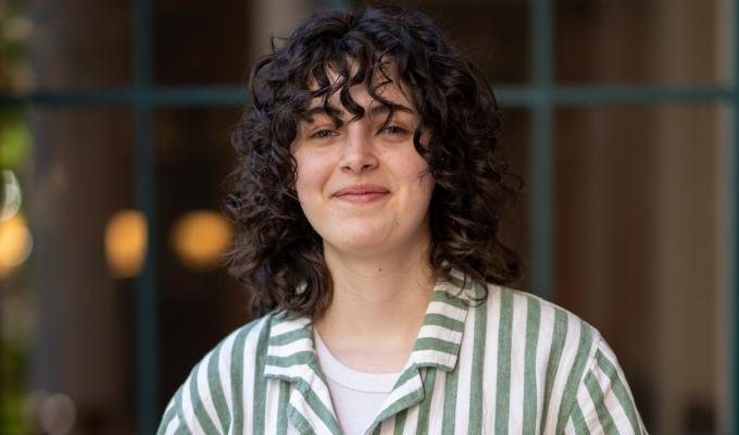 AJ Damiana wears a striped button-up with a smile, her dark, curly hair falling to her shoulders.