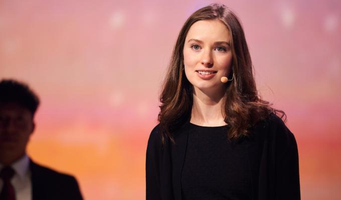 A woman with shoulder-length hair wears an earset microphone and presents to an unseen audience. Light focuses on her as stands in front of a soft orange-pink gradient.