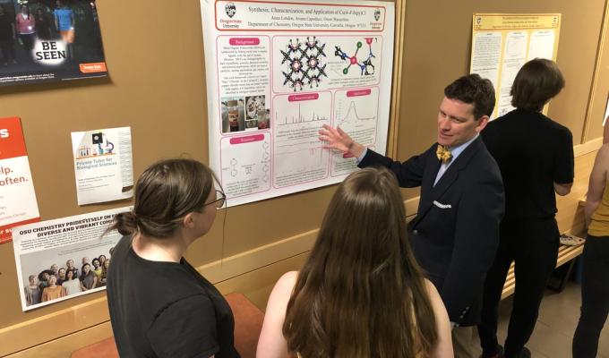 A student shows a group of people a research poster hanging on a wall.