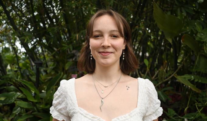 A woman in a white blouse stands in front of greenery.
