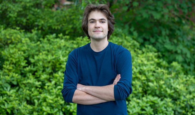 Joey Takach posing with a smile in front of leafy green bushes.