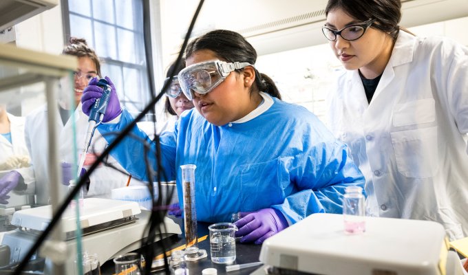 Students and staff clad in lab coats and glasses surround a lab table conducting experiments with pipettes, test tubes, beakers and other chemistry instruments in a lab at Oregon State University.