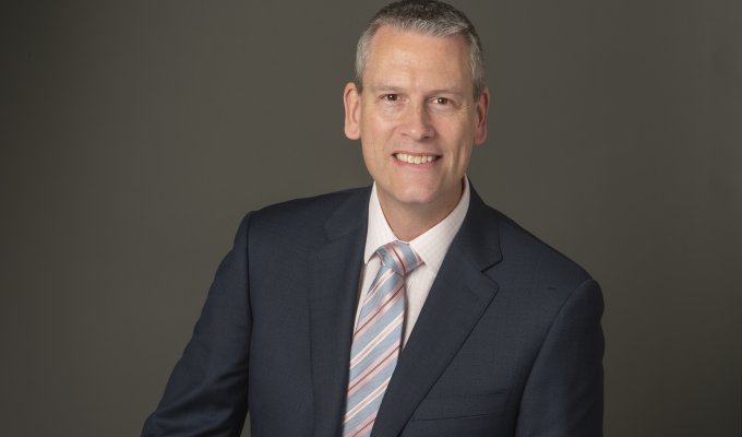 A man in a charcoal suit and striped silver tie smiles in front of a plain gray backdrop.