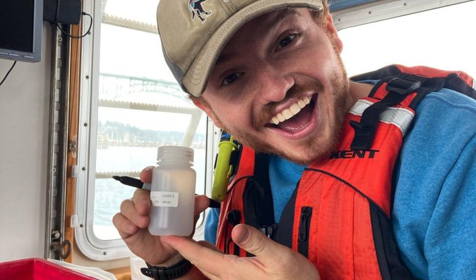 A man with a slight beard wearing a tan hat with a bison on it, and wearing an orange lifevest, holds a sample bottle.