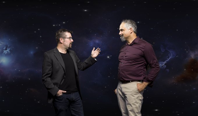 Two men stand in front of a background of space on a green screen.