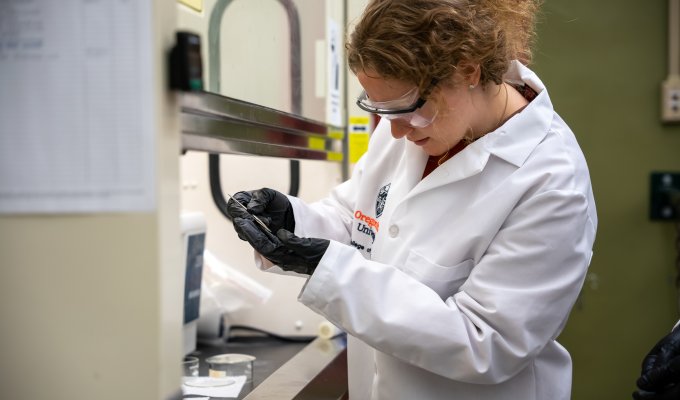 A student with her hair pulled back wearing a lab coat and protective goggles looks intently at the glassware in her hands.