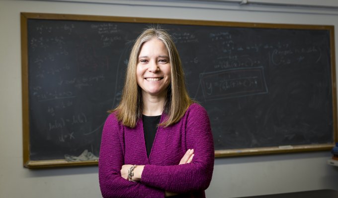 A woman in a purple suit coat stands in front of a chalkboard for a headshot.