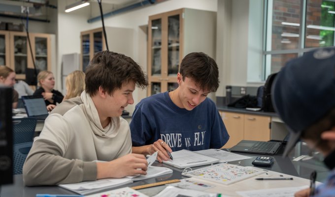 Two male students sit at a table with notebooks working in a classroom.
