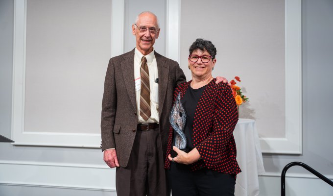 A man in a brown suit stands next to a woman wearing a red and black dress jacket. The woman is holing a glass award.
