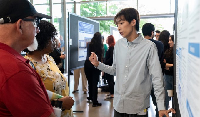 In a busy, sun-lit row lined with research posters, presenters and attendees, a young man in a button-up shirt discusses his poster with two others. 