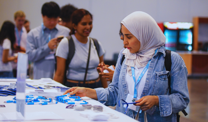 Woman wearing a hijab and a blue JSM lanyard peruses merchandise on a table with other conference attendees milling around.