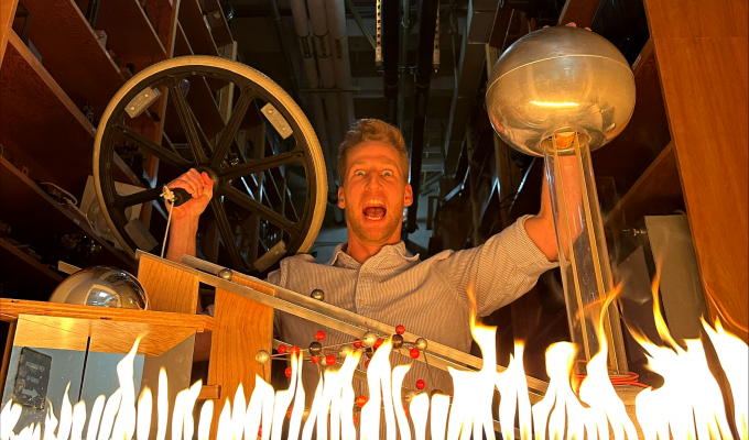 A man in a stripped shirt holds a bicycle wheel and other physics demonstration tools sit on the table in front of him and there is fire at the bottom of the photo.