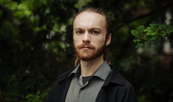 A man in a grey shirt and black coat poses for a photo in front of trees.