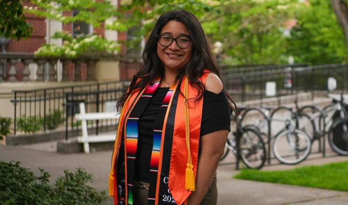 A woman in glasses and graduation regalia smiles brightly in front of a sunny campus backdrop.