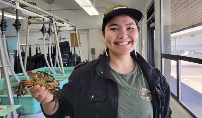 Ingram standing the Hatfield Marine Science Center wet lab, holds up a crab. She wears a CRITFIC shirt, baseball cap and beaded earrings.