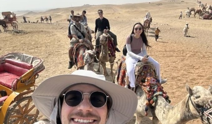 A man in a hat poses while riding a camel.
