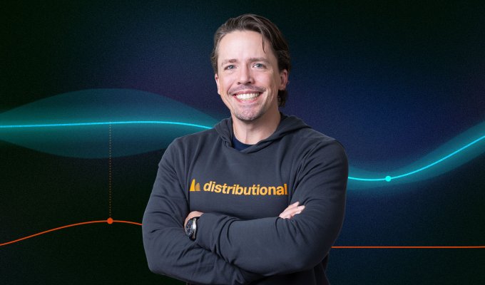 A man in a sweatshirt stands in front of a blue background.