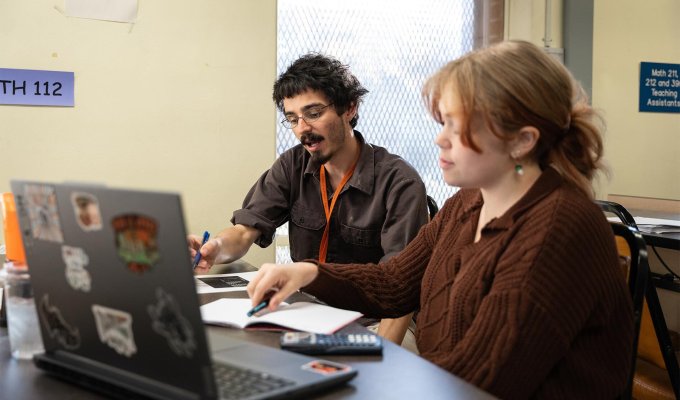 A teaching assistant sits beside a student at a table, pointing to coursework with a pen as they work together. An open laptop, notebook and calculator are on the table, and a MTH 112 sign hangs on the wall behind them.