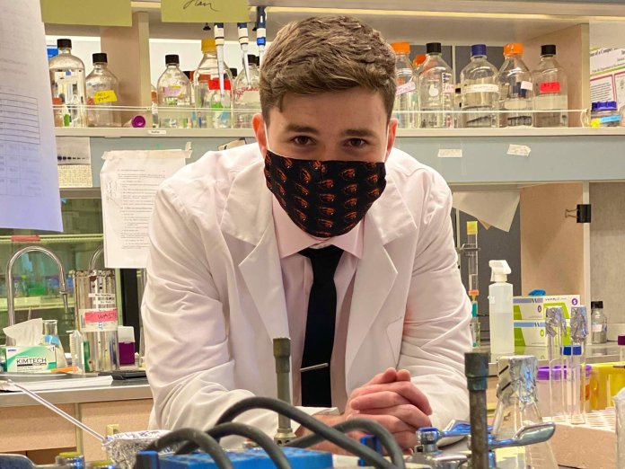 A young man in a lab coat and face mask leaning over a counter with test tubs