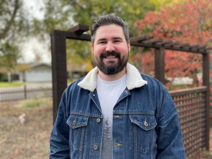 A man with a beard stands in a denim jacket with autumn colored trees in the background.