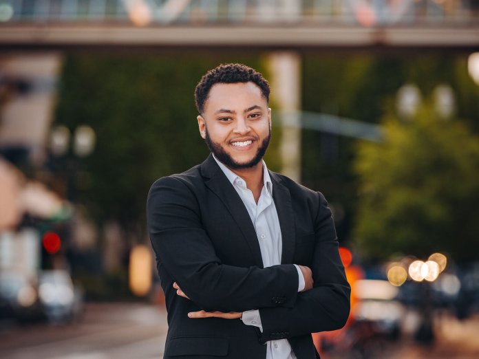 A man in a black suit stands in front of a blurry city background.