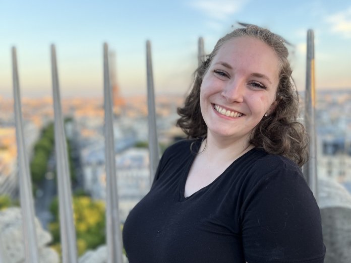A woman with a dark t-shirt and pulled back hair smiles widely at the camera, the cityscape of Paris blurred in a sunset behind her.