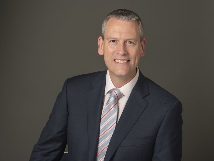 A man in a charcoal suit and striped silver tie smiles in front of a plain gray backdrop.