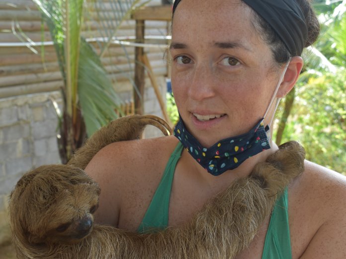 A woman in a green tank top holds a sloth in her arms, surrounded by tropical foliage.