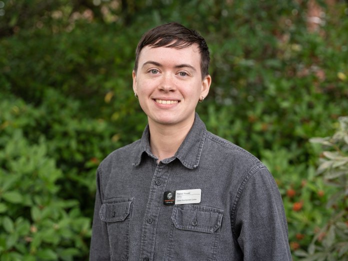 Parker smiles in front of greenery outside. They are wearing a gray button-up shirt and have short, brown hair.
