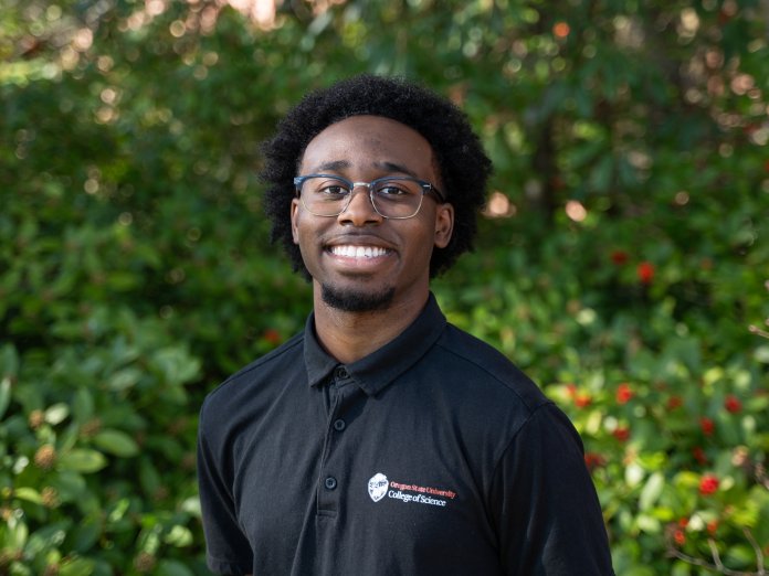 Nate smiles in front of greenery on campus. He wears glasses and a College of Science polo shirt.
