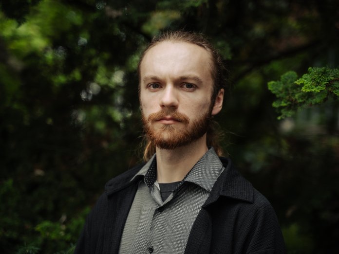 A man in a grey shirt and black coat poses for a photo in front of trees.